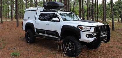 An image showing a white Toyota Tacoma in the woods. The truck has a large camper shell on the bed.