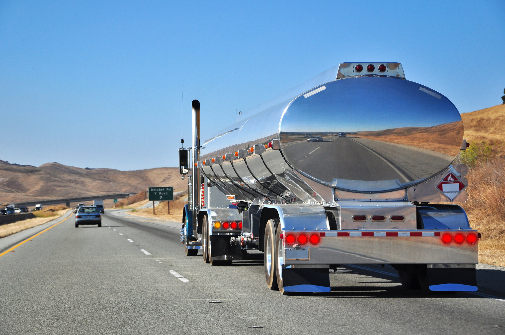 An image of multiple vehicles travelling down a highway. The focus of the image is on a larger tanker truck with rolling hills and a clear blue sky in the background.