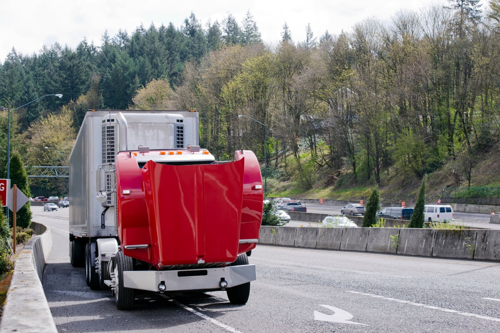 An image of a large freight truck parked alongside a roadway. The hood of the truck is open and ready to be inspected by a technician. The truck is red and pulling a silver trailer. In the backgroundd, cards blur as motorists drive by at a rapid pace.