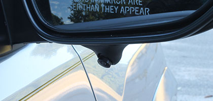 An image showing a blind spot camera attached to the bottom of a vehicle side view mirror to provide the driver with an image showing the lane beside them while the vehicle is in motion.