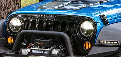 An image showing the front end of a blue Jeep Wrangler with Rostra's 7-inch LED headlamp installed and powered on.