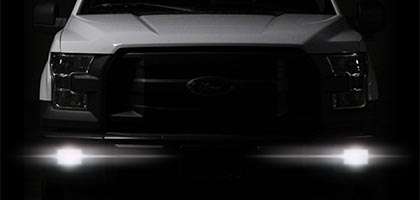 An image showing the front end of a Ford truck with Rostra LED fog lamps isntalled. The background is dark and the fog lamps are powered on illuminating the ground in front of the truck.