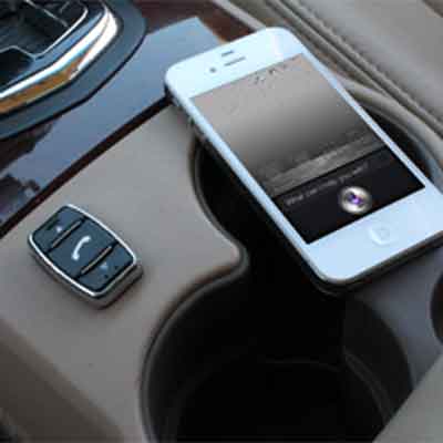 Mobile phone sitting on a vehicle center console next to the control switch for the Con-Verse hands-free Bluetooth system. The screen of the phone is illuminated showing the personal assistant Siri on an iPhone.